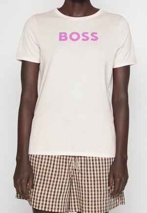 Persoon draagt een lichtroze T-shirt met de tekst "BOSS" in paarse letters en een korte broek met een bruin-wit geruit patroon.