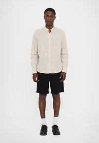 Camicia a maniche lunghe color beige chiaro con colletto cinese, righe verticali e chiusura con bottoni, abbinata a pantaloni corti neri e scarpe nere.