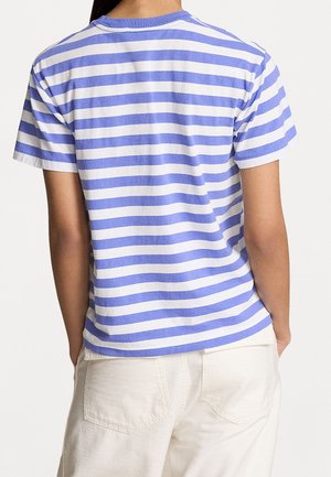 Henkilö, jolla on lyhyet hihat sinivalkoraidallinen paita ja vaaleanbeiget housut, nähtynä takaa käsien ollessa taskuissa.