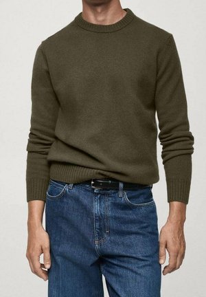 Adulte portant un pull en maille vert olive et un jean bleu avec ceinture noire, debout devant un fond clair uni.