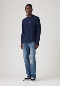 Mees, kellel on punutud juuksed, kannab tumesinist pikkade varrukatega särke, heledat sinist teksapükse ja musti tossusid, seistes ühetoalise heleda tausta vastu.