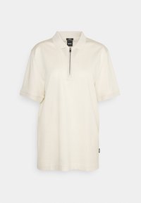 Polo tričko s krátkým rukávem v světle krémové barvě se zipovým límcem, vyrobené z měkké, texturované látky. Obsahuje malou logo nášivku na boku.