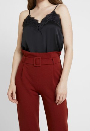 Frau trägt ein schwarzes Spitzen-Spaghettiträgertop, das in hoch taillierte, kastanienbraune Hose mit passendem Gürtel und Schnalle gesteckt ist.