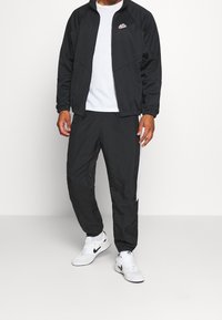 Man in zwarte Nike trainingsjack en -broek met een wit T-shirt en witte Nike sneakers tegen een effen achtergrond.