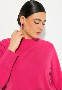 Kuum pinkki neule, jossa on korkea kaulus, ribbattu hihansuu ja sileä tekstuuri. Suuret metalliset korvakorut täydentävät ilmeen.