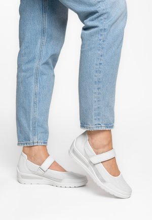 Persoon draagt lichtblauwe spijkerbroek en witte gesloten Mary Jane-schoenen met klittenbandsluitingen, staand op een witte achtergrond.