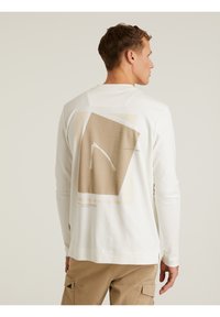 Langärmliges, weißes Baumwoll-T-Shirt mit einem geometrischen beigen Grafikdesign auf der Rückseite, einschließlich dezentem Text und einem lockeren Schnitt.