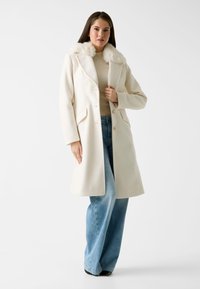 Frau in einem weißen langen Mantel mit Kunstfellkragen, beigem Pullover, hellblauen weit geschnittenen Jeans und schwarzen spitzen Schuhen vor weißem Hintergrund.