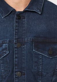 Tummansininen farkkutakki, jossa on viistot kaulukset, kaksi rintataskua ja mustat napit. Materiaali on pehmeää ja siinä on näkyviä kudontakuvioita.