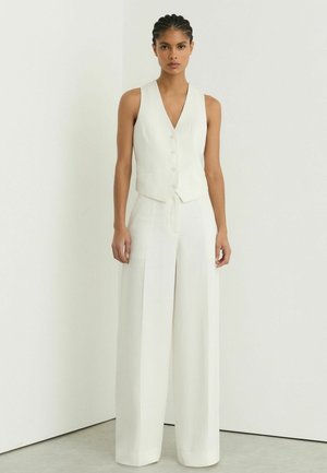 Naine seisab valge taustaga, kandes valget varrukateta nööbitavat vesti ja laiade säärtega valgeid pükse.