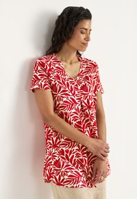 Rød og creme kortærmet top med et blomstret bladmønster, v-hals og afslappet pasform. Stoffet virker let og blødt.