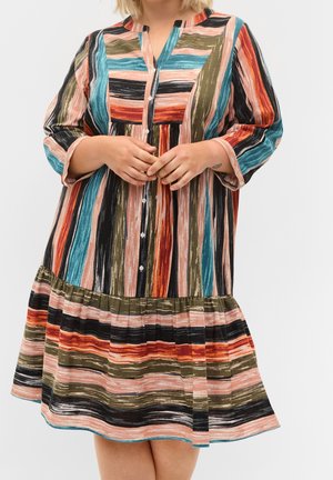 Frau trägt ein langärmliges, knöpfbares Kleid mit einem mehrfarbigen, vertikalen und horizontalen Streifenmuster und einem gekräuselten Saum.