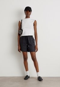 Top bianco senza maniche, pantaloni corti neri con coulisse, cappellino nero e mocassini con calzini bianchi. Texture liscia, vestibilità casual, design minimalista.