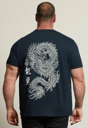 Mann trägt schwarzes T-Shirt mit detailliertem weißen Drachenmotiv und zwei vertikalen chinesischen Schriftzeichen auf dem Rücken, vom Kamera weggewandt.