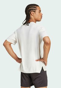 Weißes T-Shirt mit kurzen Ärmeln und geradem Schnitt, mit seitlichen Schlitzen am Saum, kombiniert mit dunklen Shorts. Glatte Textur, minimalistisches Design.