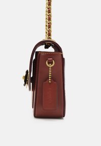 Borsa a tracolla in pelle marrone con catena dorata e una targhetta pendente con il nome del marchio, mostrata di lato su uno sfondo bianco.