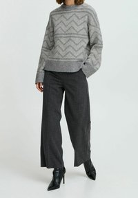 Femme portant un pull à motif zigzag gris, un pantalon large sombre et des bottes noires pointues à talons hauts, debout avec une main dans la poche.