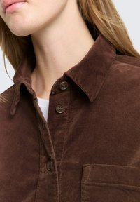 Bruine corduroy overhemd met een klassieke kraag, twee knopen en een borstzak. Stof heeft een zachte textuur met een lichte glans.