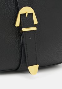 Musta nahkainen laukkuremmissä teksturoitu pinta, jossa kullanvärinen metallisolki, joka on kaartuva, sekä kolmiomainen kullanvärinen koriste remmin päässä.