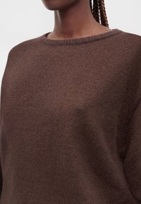 Gros plan d'une personne portant un pull en laine marron à col rond, cheveux tressés visibles sur l'épaule.