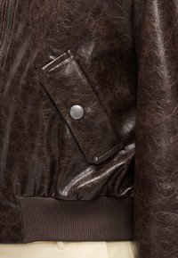 Brown faux leather bomber jacket with a glossy texture, featuring a snap pocket and ribbed hem for added detail and contrast.
