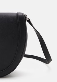 Czarna skórzana torebka na ramię o półkolistym kształcie, teksturowanej powierzchni i cienkim, płaskim pasku. Posiada srebrzysty zamek błyskawiczny jako detal.