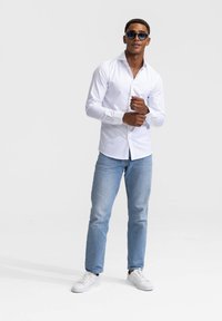 Witte blouse met lange mouwen, knoopjes, met een gladde textuur. Gecombineerd met lichtblauwe jeans en witte sneakers. Zonnebril maakt de look compleet.