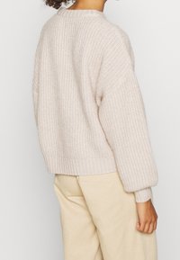 Personne portant un pull en maille épaisse couleur crème à manches longues et un pantalon beige, vue de dos sur un fond uni.