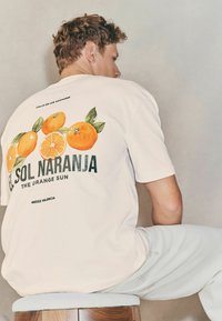 Maglietta bianca in cotone con illustrazioni arancioni vivaci e testo verde che recita "EL SOL NARANJA THE ORANGE SUN", con una vestibilità casual.