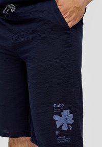 Navyblauwe gestructureerde shorts met zakken. Kenmerkt een blauw bloemenlogo en tekst op het onderbeen. Verstelbare drawstring tailleband.
