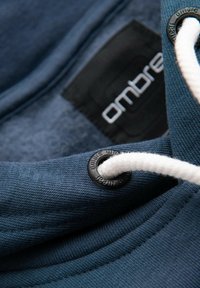 Sweat à capuche bleu marine avec intérieur en peluche, cordon blanc à ajuster, et une étiquette noire avec le logo "ombre".
