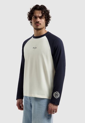 Man met krullend haar die een wit en marineblauw raglanshirt met lange mouwen draagt, met een tekst op de borst en een embleem op de mouw, gecombineerd met lichtblauwe spijkerbroek.