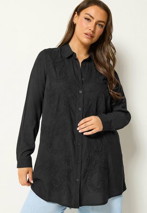 Kvinde med langt brunt hår iført en sort skjorte med knapper og diskret blomsterbroderi samt lyseblå jeans, stående mod en lys baggrund.