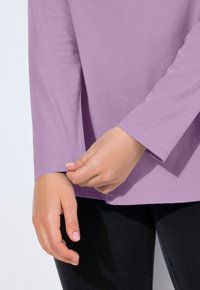 Ruce upravují manžetu dlouhého lila rukávu košile na pozadí s tmavými kalhotami na světlém jednobarevném pozadí.