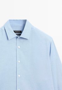 Světle modrá košile s knoflíky z texturované látky má klasický límec a bílé knoflíky, štítek viditelný uvnitř límce.
