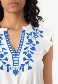 Λευκό μπλουζάκι με μπλε λουλουδάτο κέντημα, V-λαιμόκοψη και βολάν μανίκια. Διαθέτει περίπλοκο ράψιμο και υφή υφάσματος.