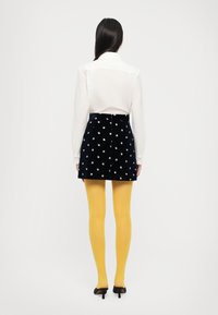 Granatowa aksamitna spódnica A-line z kwiatowym haftem, zestawiona z żółtymi, nieprzezroczystymi rajstopami i białą koszulą z zapięciem na guziki. Czarne obcasy.