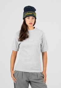 Giovane donna che indossa una maglietta grigia a maniche corte, pantaloni grigi plissettati e un berretto nero con testo giallo su uno sfondo chiaro semplice.