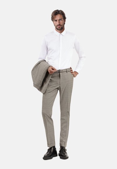 Hellgraue karierten Hosen, kombiniert mit einem weißen Hemd und einem grauen Blazer. Mann steht selbstbewusst, schwarze Schuhe sind sichtbar.