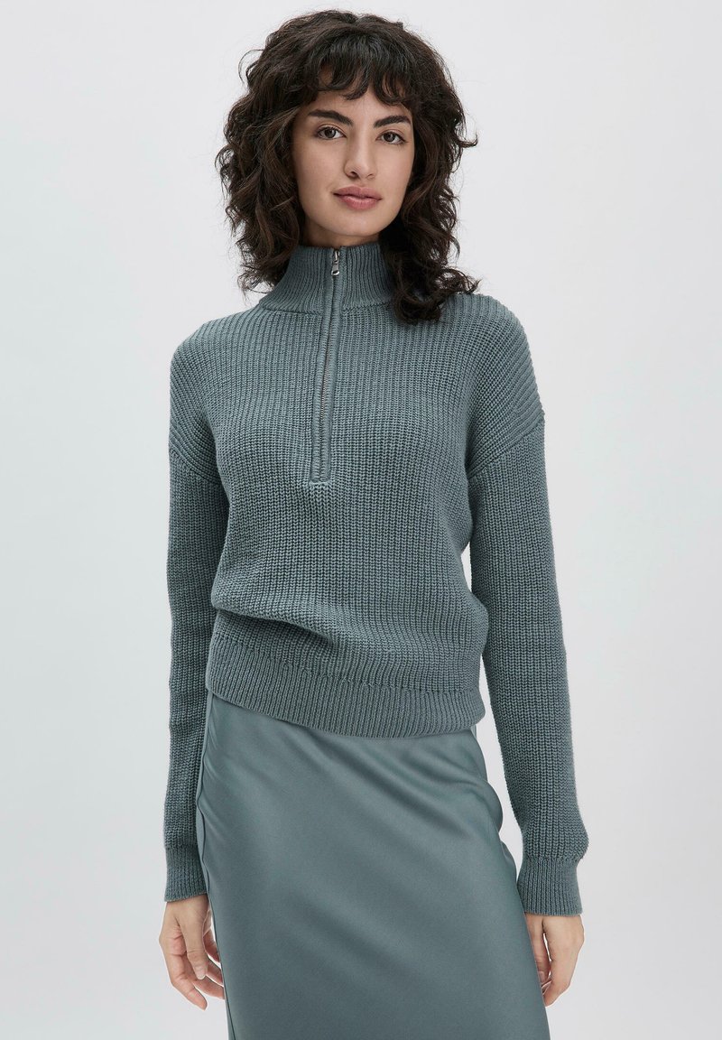 Pull en maille grise à col montant et fermeture éclair quart, avec des poignets côtelés et un motif texturé, associé à une jupe grise lisse.