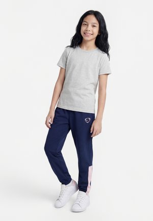 Jong meisje met lang donker haar, draagt een grijs T-shirt, marineblauwe broek met roze accenten en witte sneakers, staand op een witte achtergrond.