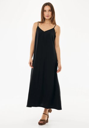 Kobieta ubrana w czarną, bezrękawnikową sukienkę maxi i brązowe sandały na wysokim obcasie z panterkowym wzorem, stojąca na tle jednolitego, białego tła.