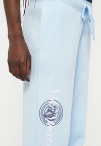 Svetlo modre trenirke z mehko teksturo, ki imajo velik okrogli logotip in napis "Nike Sports" v beli barvi, poudarjene z vrvico za prilagajanje v pasu.