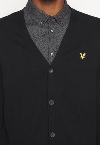 Schwarze gestrickte Strickjacke mit V-Ausschnitt, mit fünf Knöpfen und einem kleinen gelben Adler-Logo. Über einem grauen Hemd mit Knöpfen getragen.