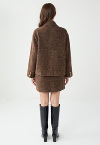 Braune Tweedjacke mit Kragen und langen Ärmeln, kombiniert mit einem passenden Minirock. Schwarze kniehohe Stiefel. Glattes, strukturiertes Gewebe.