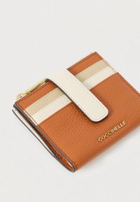 Compact leather wallet in tan and cream with a snap strap and gold Coccinelle logo, displayed on a white background.