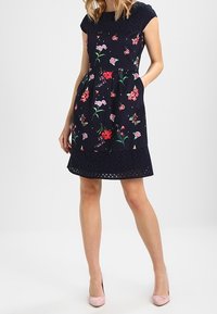 Abito floreale blu navy a maniche corte, con corpetto aderente, tasche e orlo decorato con occhielli. Indossato con scarpe rosa.
