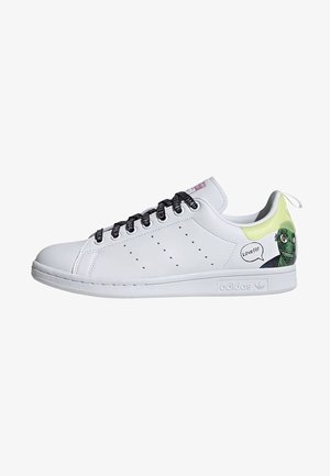 Baskets blanches Adidas avec lacets noirs, bandes latérales perforées et visage de dessin animé vert avec bulle de dialogue au talon.