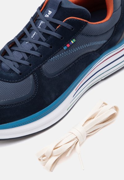 Αθλητικό παπούτσι σε ναυτικό μπλε χρώμα με πορτοκαλί εσωτερική επένδυση, πολύχρωμες ρίγες στη λ ευκή σόλα και επιπλέον κορδόνια σε μπεζ απόχρωση δίπλα του.