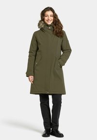 Olijfgroene, knielange parka met een met bont gevoerde capuchon, drukknoopsluitingen en zijzakken, gedragen over zwarte broek en enkellaarsjes.